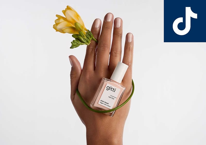 Hände ineinandergelegt, eine Hand hält einen gelben Blumenstängel, um die Hand gewickelt eine transparente Nagellack-Flasche mit weißer Kappe und dem Schriftzug „glitt“ auf dem Etikett, oben rechts kleines weißes TikTok-Logo auf dunkelblauem Quadrat