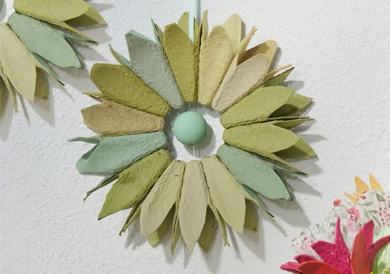 Kranz aus etwa 20 Papierblättern in verschiedenen Grüntönen und Beige, die sternförmig um einen kleinen runden, hellgrünen Mittelpunkt angeordnet sind und an einer Wand hängen