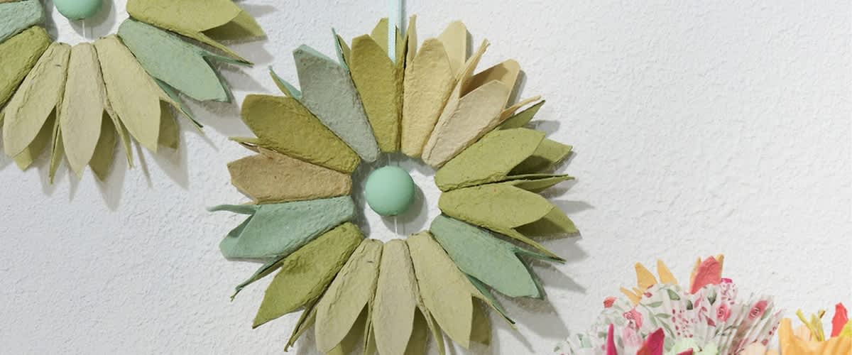 Kranz aus etwa 20 Papierblättern in verschiedenen Grüntönen und Beige, die sternförmig um einen kleinen runden, hellgrünen Mittelpunkt angeordnet sind und an einer Wand hängen