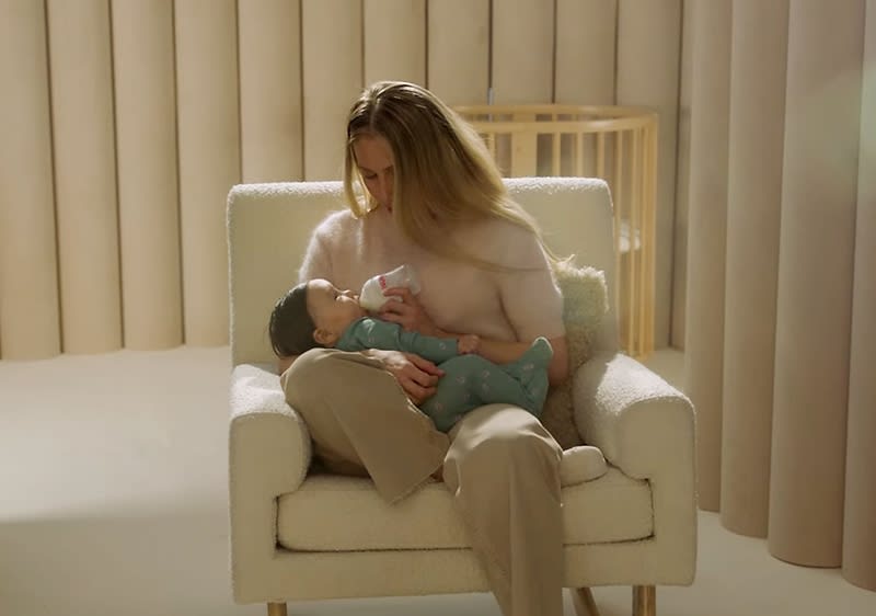 Eine sitzende Erwachsene in heller Kleidung stillt ein Baby im Arm, das in einem grünen Strampelanzug liegt, auf einem cremefarbenen Sessel vor beigen Vorhängen und einem Holzgitter als Deko im Hintergrund