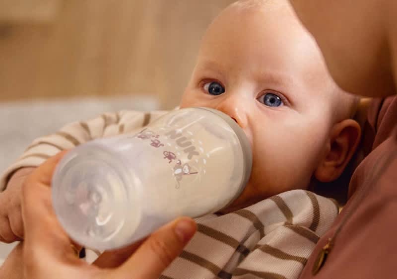 Eine Person hält ein Baby im Arm, das an einer transparenten Babyflasche mit weißem Sauger trinkt; Baby trägt ein langärmeliges, gestreiftes Schlafanzugoberteil und die betreuende Person trägt ein rosafarbenes Oberteil