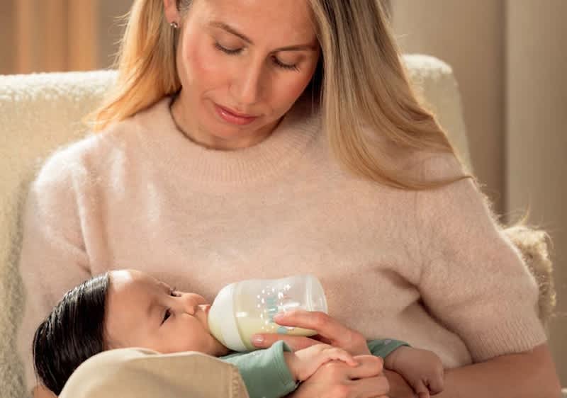 Eine Frau sitzt in einem Sessel und füttert ein Baby mit einer Milchflasche, die sie mit beiden Händen hält, während das Baby in ihren Armen liegt und an der Flasche saugt