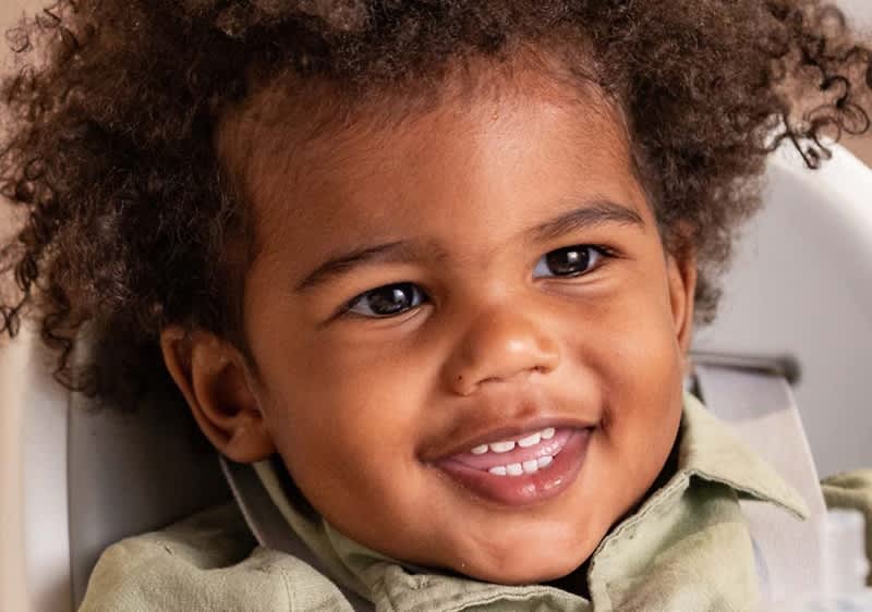 Kleines Kind mit lockigem Haar, sitzt in einem Kindersitz und trägt ein hellgrünes Hemd, Gesicht unkenntlich gemacht durch zentrierten Farbverlauf, nur Ohr und Haare seitlich sichtbar