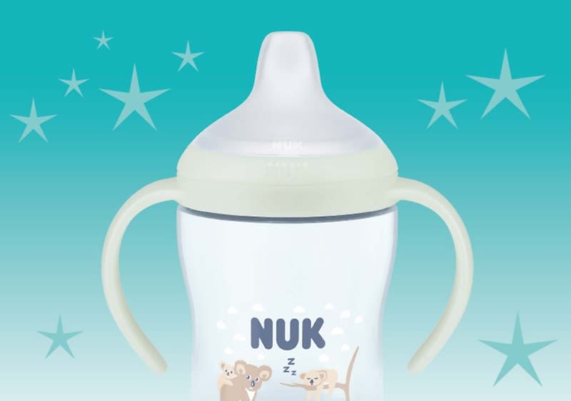 Ein transparenter NUK-Trinklernbecher mit zwei seitlichen Griffen, hellem Saugerdeckel und Aufdruck mit zwei schlafenden Tierfiguren und dem Schriftzug „NUK“ vor türkisem Hintergrund mit Sternen