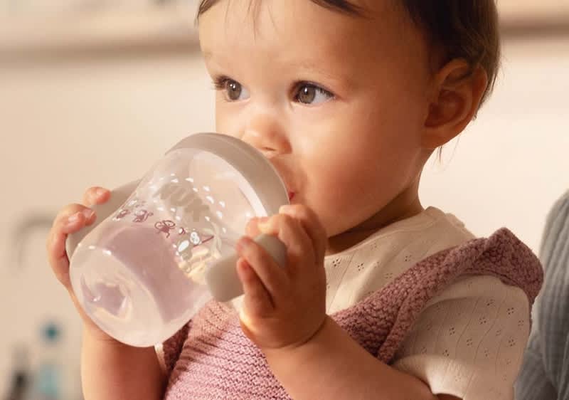 Kleines Kind sitzt aufrecht und trinkt aus einem durchsichtigen Babybecher mit grauem Deckel, trägt ein weißes Oberteil und ein rosa Stricktäschchen über der Brust, blickt nach rechts und hält den Becher mit beiden Händen
