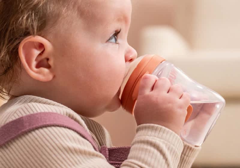 Kleinkind im Profil, das aus einem transparenten Babyfläschchen mit orangefarbenem Verschluss trinkt, die Hände halten das Fläschchen dicht am Mund, heller Hintergrund