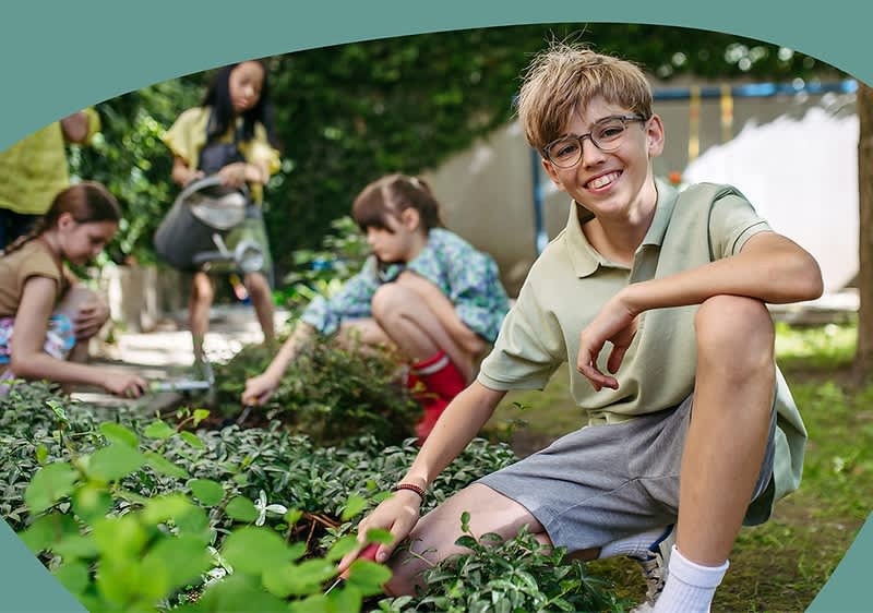 Kinder knien und hocken in einem Gartenbeet und jäten Unkraut; im Vordergrund sitzt ein Junge mit hellem T-Shirt und grauen Shorts und greift zur Erde, im Hintergrund arbeiten zwei weitere Kinder am Beet und ein weiteres Kind schiebt eine Schubkarre, alle in sommerlicher Freizeitkleidung und umgeben von grünem Pflanzenbewuchs