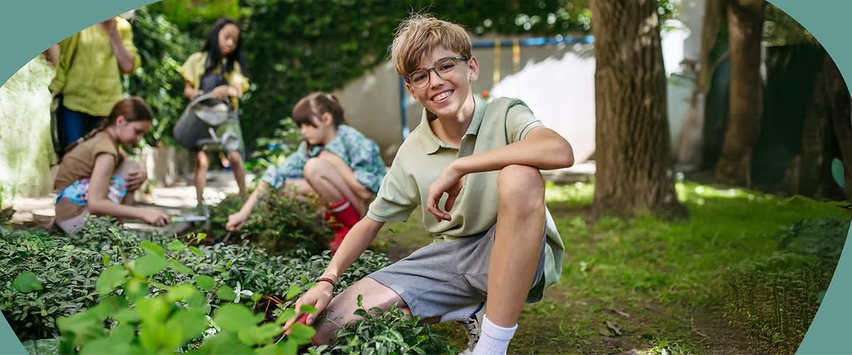 Kinder knien und hocken in einem Gartenbeet und jäten Unkraut; im Vordergrund sitzt ein Junge mit hellem T-Shirt und grauen Shorts und greift zur Erde, im Hintergrund arbeiten zwei weitere Kinder am Beet und ein weiteres Kind schiebt eine Schubkarre, alle in sommerlicher Freizeitkleidung und umgeben von grünem Pflanzenbewuchs