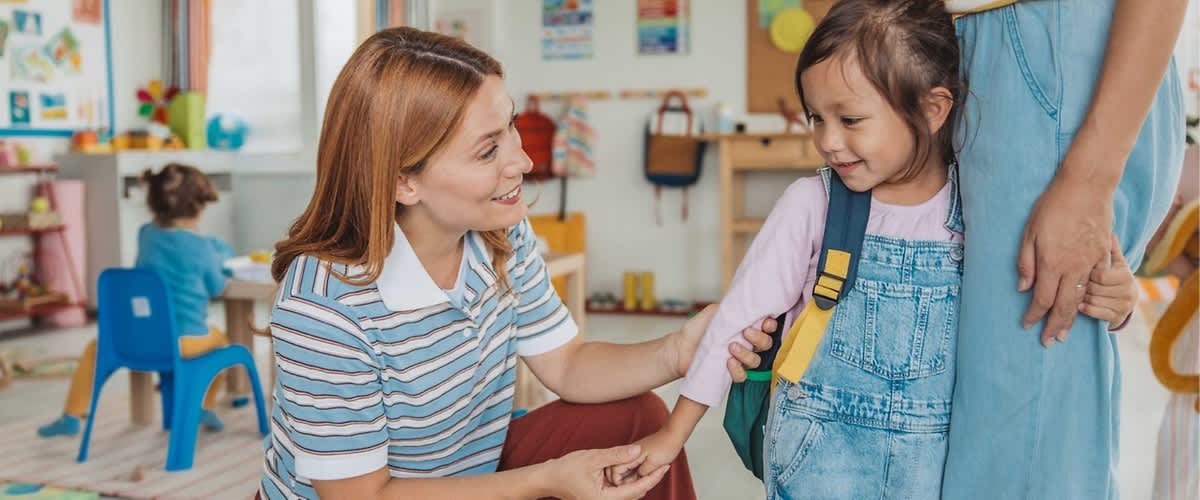 Eine Erzieherin kniet auf Augenhöhe und nimmt ein Mädchen mit Rucksack behutsam an der Hand, im Hintergrund weitere Kinder und bunte Kindergarten-Dekorationen