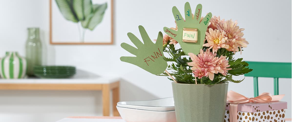 Grüner Blumentopf mit rosa Chrysanthemen, in denen drei grüne Papierhände gesteckt sind; auf einer Hand steht groß „FINN“ in roter Schrift, auf einer anderen sind verschiedene Namen in bunten Stiften geschrieben und ein kleiner Zettel mit „FINN“ befestigt; rechts neben dem Topf eine rosa Geschenkbox mit goldenen Punkten und Schleife