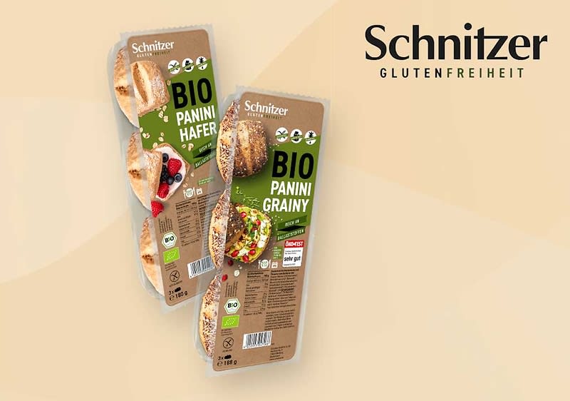 Zwei längliche braune Verpackungen des glutenfreien Produkts Schnitzer BIO Panini mit aufgedruckten Bildern von belegten Panini, grünen Etiketten und Logos, auf beigefarbenem Hintergrund rechts oben das Schriftzug-Logo Schnitzer GLUTENFREIHEIT