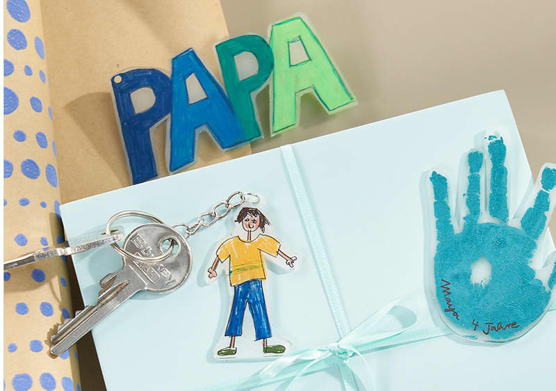Auf einem hellblauen Geschenkordner liegt eine kleine Schlüsselanhänger-Kette mit zwei Hausschlüssel, daneben ein ausgeschnittener Papiermensch mit gelbem T‑Shirt und blauer Hose sowie ein türkiser Handabdruck mit der Beschriftung „Mama & Jaune“; im Hintergrund steht aus bunten Buchstaben das Wort „PAPA“ und links ist braunes Geschenkpapier mit blauen Punkten sichtbar