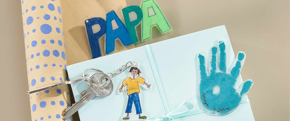 Auf einem hellblauen Geschenkordner liegt eine kleine Schlüsselanhänger-Kette mit zwei Hausschlüssel, daneben ein ausgeschnittener Papiermensch mit gelbem T‑Shirt und blauer Hose sowie ein türkiser Handabdruck mit der Beschriftung „Mama & Jaune“; im Hintergrund steht aus bunten Buchstaben das Wort „PAPA“ und links ist braunes Geschenkpapier mit blauen Punkten sichtbar