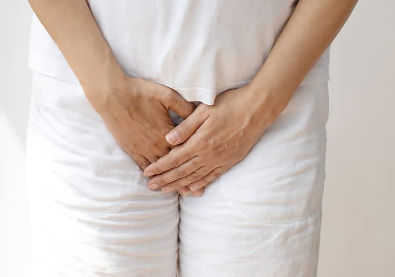 Hände einer stehenden Person liegen über dem Intimbereich, die Fingerspitzen leicht verschränkt, bekleidet mit weißem T-Shirt und weißen Hosen