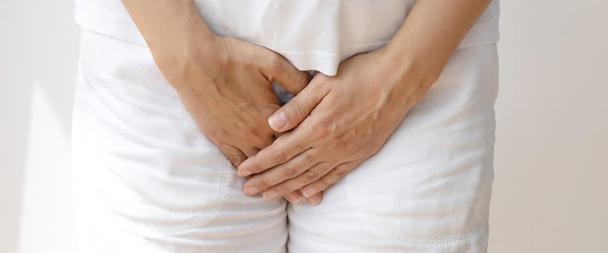 Hände einer stehenden Person liegen über dem Intimbereich, die Fingerspitzen leicht verschränkt, bekleidet mit weißem T-Shirt und weißen Hosen