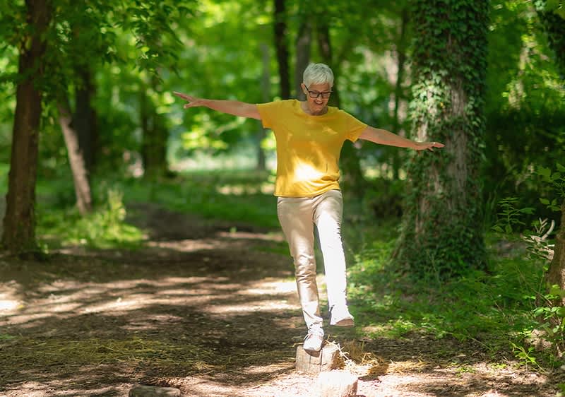 Ältere Person mit kurzem weißen Haar balanciert mit ausgestreckten Armen auf einem schmalen Baumstamm auf einem Waldweg, trägt gelbes T-Shirt und helle Hosen, sonnendurchfluteter Laubwald im Hintergrund