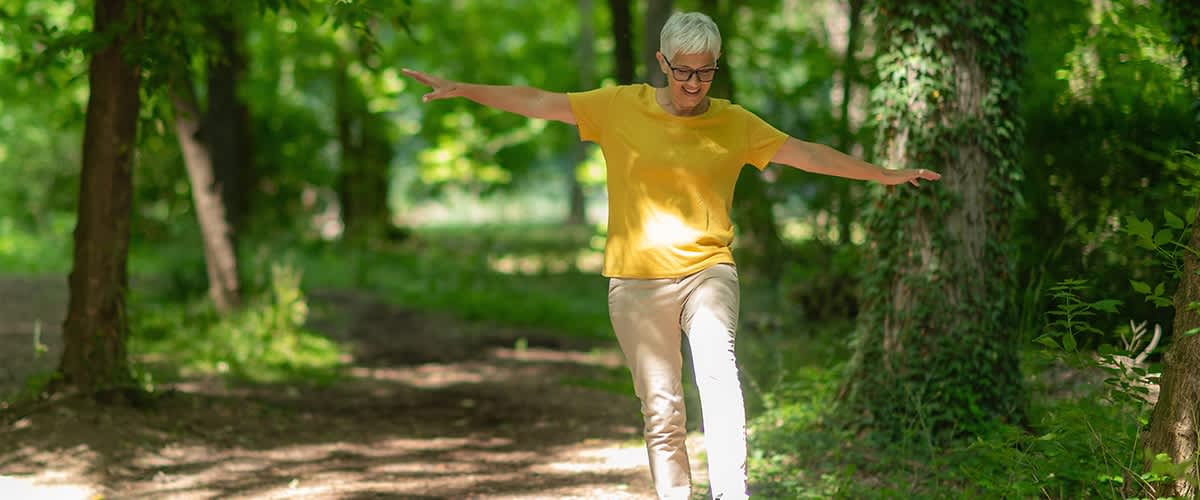 Ältere Person mit kurzem weißen Haar balanciert mit ausgestreckten Armen auf einem schmalen Baumstamm auf einem Waldweg, trägt gelbes T-Shirt und helle Hosen, sonnendurchfluteter Laubwald im Hintergrund