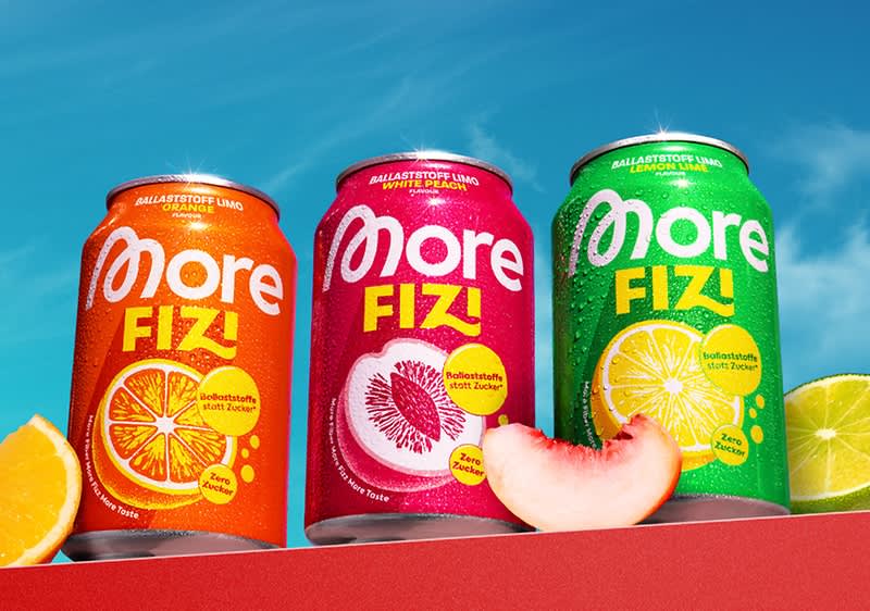 Drei bunte Getränkedosen der Marke „More Fizi“ in Reihe vor blauem Himmel: links orange Dose mit Orangenscheiben und Aufschrift „More Fizi“, mittig pinke Dose mit Grapefruitgrafik und Schriftzug, rechts grüne Dose mit Zitronenscheibe und Schriftzug, davor Fruchtstücke (Orange, Pfirsich, Zitrone) auf rotem Untergrund