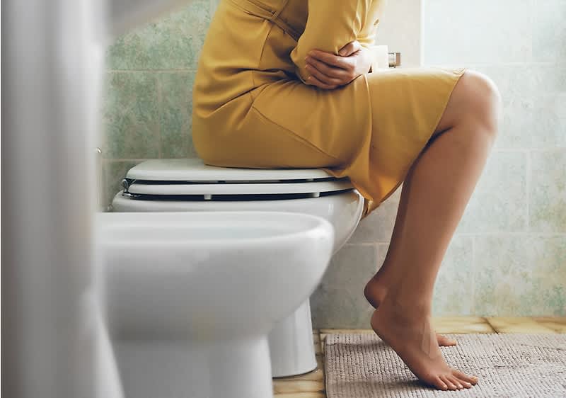 Frau im goldgelben Kleid sitzt auf der geschlossenen Toilettenbrille, die Arme verschränkt vor dem Bauch, ein Bein angewinkelt und nackte Füße auf einem Badezimmerteppich stehend