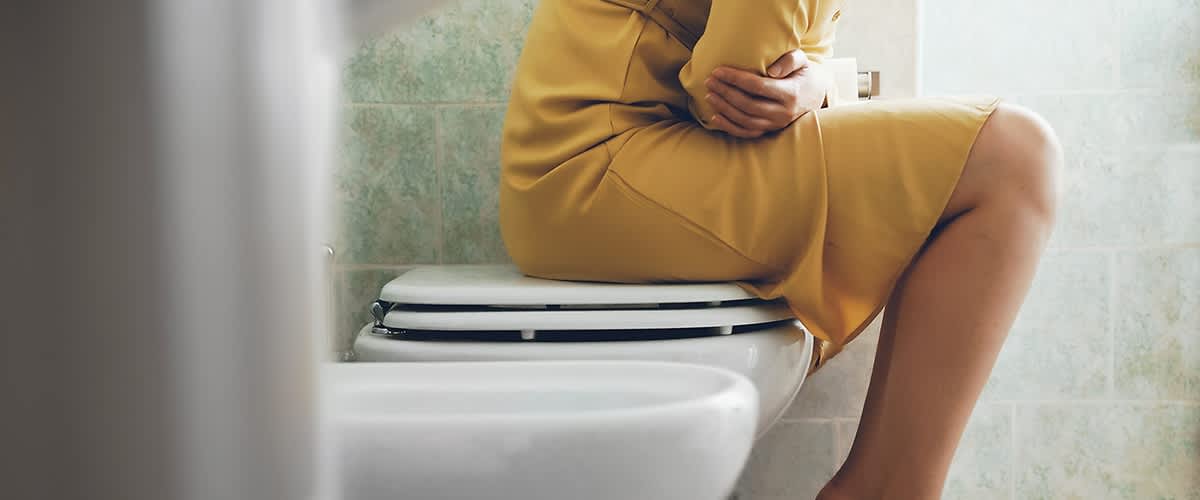 Frau im goldgelben Kleid sitzt auf der geschlossenen Toilettenbrille, die Arme verschränkt vor dem Bauch, ein Bein angewinkelt und nackte Füße auf einem Badezimmerteppich stehend