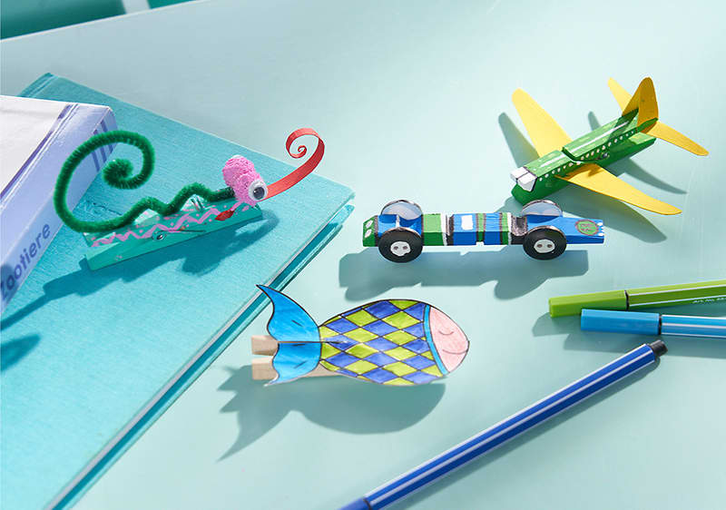 Mehrere bunte Papp- und Bastelspielzeuge auf hellblauem Untergrund: oben rechts ein grünes und gelbes Papierflugzeug, daneben ein kleines blau-grünes Rennwagen-Modell mit schwarzen Rädern, links unten ein bunt gemustertes Pappfischchen, oben links eine grüne Pfeifenputzer-Bastelspirale auf einem kleinen Pappsteg und rechts mehrere blaue und grüne Stifte nebeneinander
