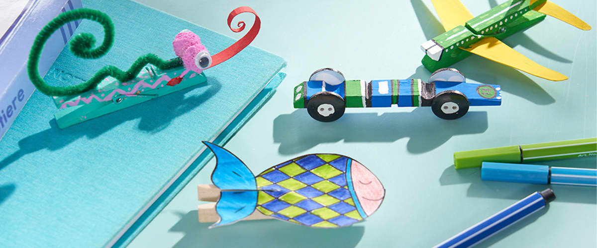 Mehrere bunte Papp- und Bastelspielzeuge auf hellblauem Untergrund: oben rechts ein grünes und gelbes Papierflugzeug, daneben ein kleines blau-grünes Rennwagen-Modell mit schwarzen Rädern, links unten ein bunt gemustertes Pappfischchen, oben links eine grüne Pfeifenputzer-Bastelspirale auf einem kleinen Pappsteg und rechts mehrere blaue und grüne Stifte nebeneinander