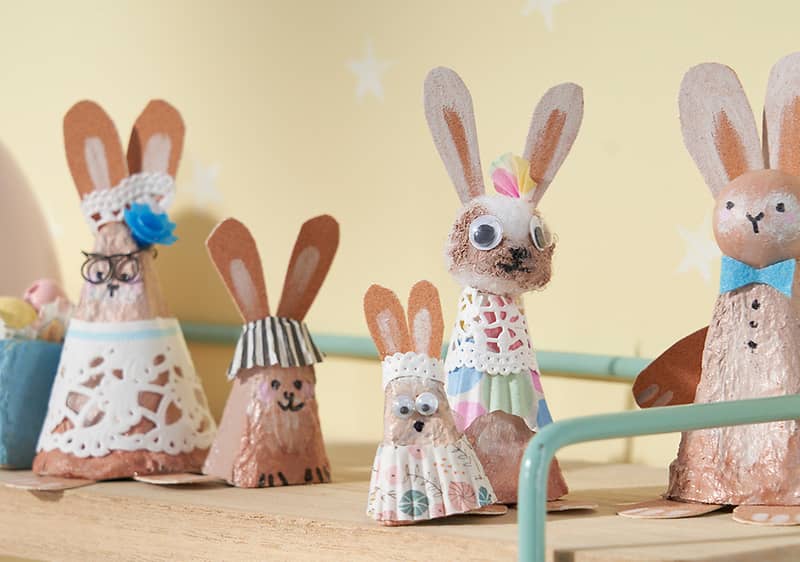Sechs handgefertigte Osterhasen aus bemalten Eierschalen und Papier, auf einem Holzbrett arrangiert; Vordergrund: kleiner Hase mit Perlenaugen und gepunktetem Papierrock, daneben ein Hasenkopf mit Papierohren und Spitzenband als Haarschmuck; Mitte: zwei Häschen mit Papierohren, eines mit Papierschürze und ein größeres mit einer Wackelauge und gehäkeltem Kragen; rechts ein größerer Hase mit blauem Papierfliegenband und kupferfarbenem Körper; hellgelber Hintergrund mit weißen Sternen