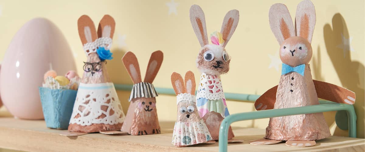 Sechs handgefertigte Osterhasen aus bemalten Eierschalen und Papier, auf einem Holzbrett arrangiert; Vordergrund: kleiner Hase mit Perlenaugen und gepunktetem Papierrock, daneben ein Hasenkopf mit Papierohren und Spitzenband als Haarschmuck; Mitte: zwei Häschen mit Papierohren, eines mit Papierschürze und ein größeres mit einer Wackelauge und gehäkeltem Kragen; rechts ein größerer Hase mit blauem Papierfliegenband und kupferfarbenem Körper; hellgelber Hintergrund mit weißen Sternen