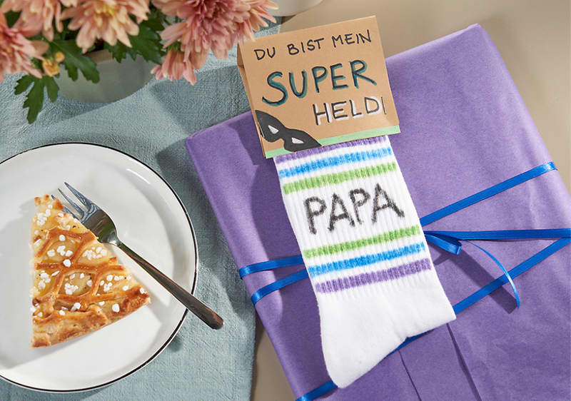 Auf einem hellen Tisch liegt ein lila Geschenkpaket mit blauem Band, darauf ein Paar weiße Kindersocken mit grünen und blauen Streifen und dem Aufdruck „PAPA“, in den Socken steckt eine kleine Karte mit der handschriftlichen Aufschrift „DU BIST MEIN SUPER HELD“, daneben ein weißer Teller mit einem Stück Kuchen und einer Gabel sowie rosa Blumen am oberen Bildrand