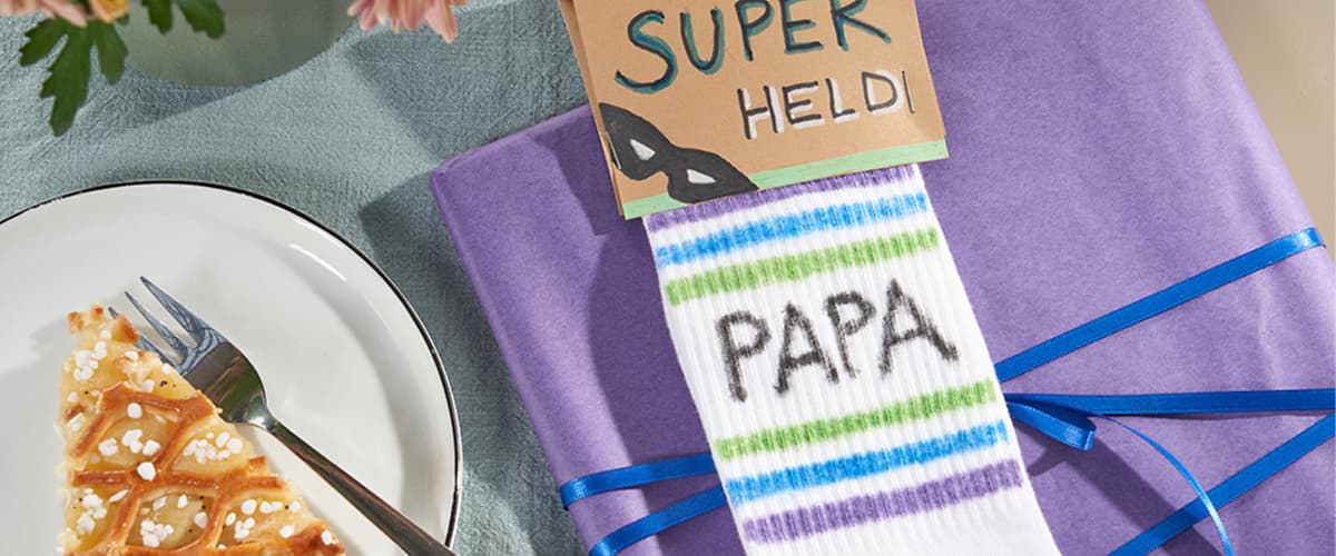 Auf einem hellen Tisch liegt ein lila Geschenkpaket mit blauem Band, darauf ein Paar weiße Kindersocken mit grünen und blauen Streifen und dem Aufdruck „PAPA“, in den Socken steckt eine kleine Karte mit der handschriftlichen Aufschrift „DU BIST MEIN SUPER HELD“, daneben ein weißer Teller mit einem Stück Kuchen und einer Gabel sowie rosa Blumen am oberen Bildrand
