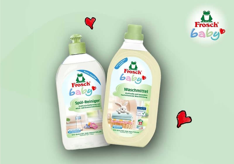 Zwei Frosch Baby Reinigungsprodukte vor hellgrünem Hintergrund: links eine weiße Flasche mit grünem Klappverschluss und Aufschrift „Frosch baby Spül-Reiniger“ mit Babyflaschen-Illustration, rechts eine beige-flaschenförmige Packung mit grünem Schraubverschluss und Aufschrift „Frosch baby Waschmittel“ mit Wäsche- und Plüschtier-Illustration, oben rechts das Frosch-Logo und drei kleine rote Herz-Illustrationen