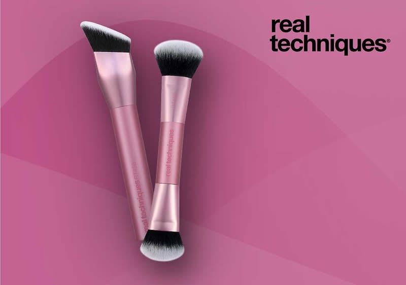 Zwei rosa Make-up-Pinsel der Marke Real Techniques mit schwarzen und weißen Borsten liegen überkreuzt auf rosa Hintergrund, rechts oben das schwarze Logo „real techniques“