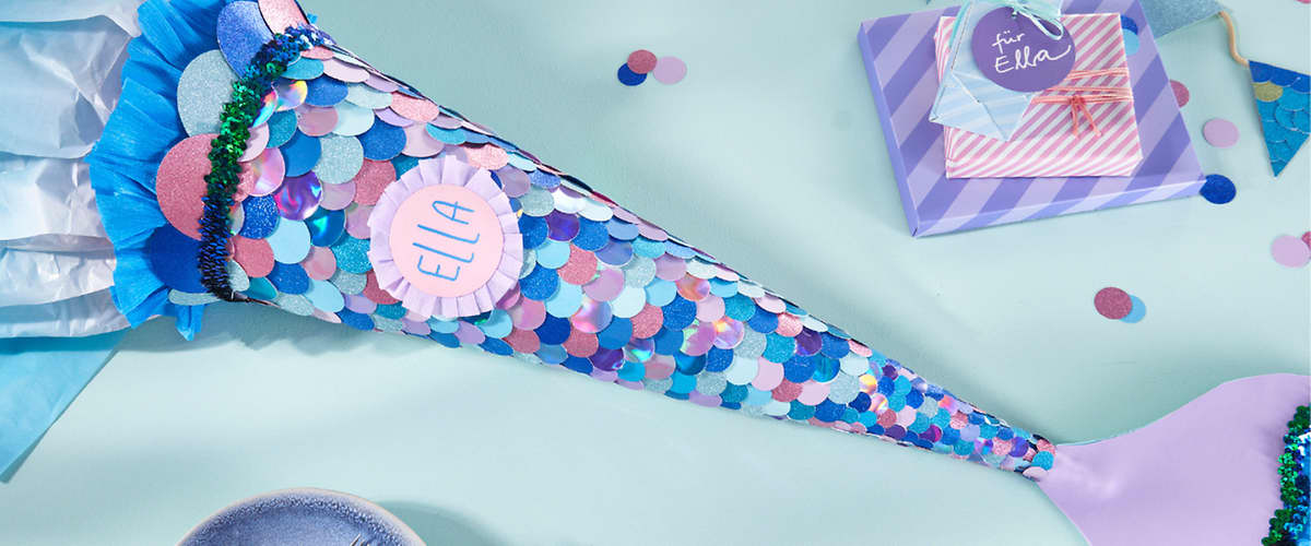 Längliche Schultüte mit schillernden Pailletten in Blau-, Pink- und Lilatönen, am oberen Ende mit grünem Fransenabschluss und rundem Namensschild „Ella“, auf hellgrünem Untergrund neben blau-lila Geschenkbox mit Anhänger „für Ella“ und Partykonfetti, im Vordergrund Teller und Besteck sichtbar