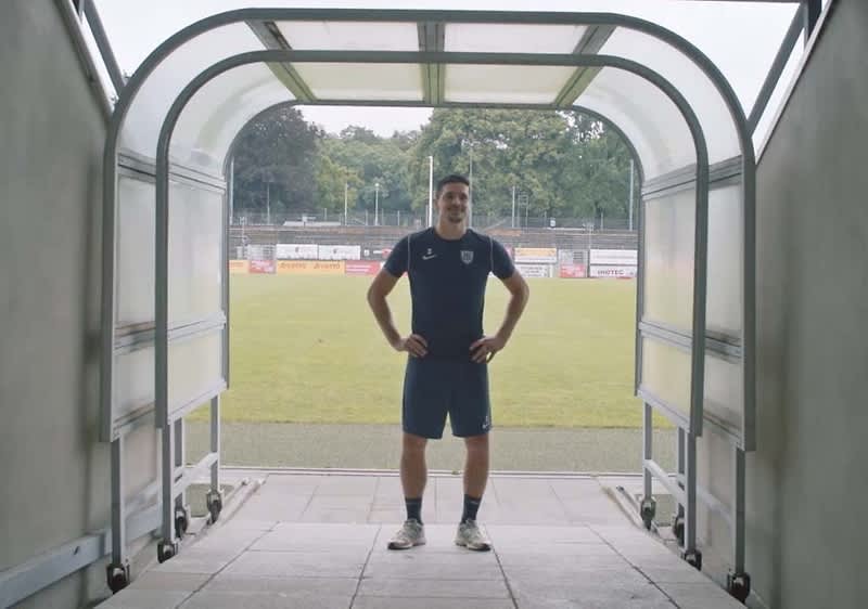 Ein junger Mann in dunkelblauem Sporttrikot und kurzen Hosen steht mit den Händen in die Hüften am Ausgang eines halbtransparenten Spielertunnels und blickt auf ein leeres Fußballfeld hinter ihm