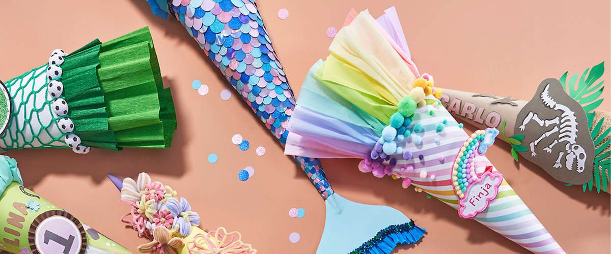 Bunte Partyhüte und -hörner auf rosafarbenem Untergrund, darunter ein hornförmiger Hut mit blauen Glitzerschuppen, ein Papiertütenhut in Regenbogenfarben mit Pompons und Aufkleber „Fina“, ein pastellgelber Hut mit Schleifen und Blumenverzierungen sowie verstreutes rundes Konfetti