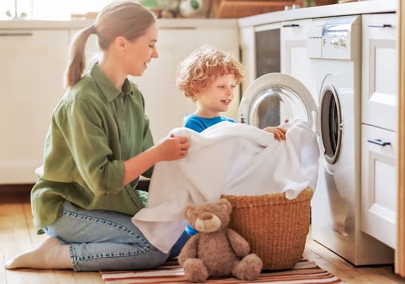 Zwei Personen, eine ältere sitzend im Schneidersitz und eine jüngere auf den Knien, legen zusammen weiße Wäsche aus einem Weidenkorb in die Frontlader-Waschmaschine der Küche; im Vordergrund liegt ein brauner Teddybär auf einem gestreiften Teppich, im Hintergrund Küchenschränke und Waschmaschinentür sichtbar
