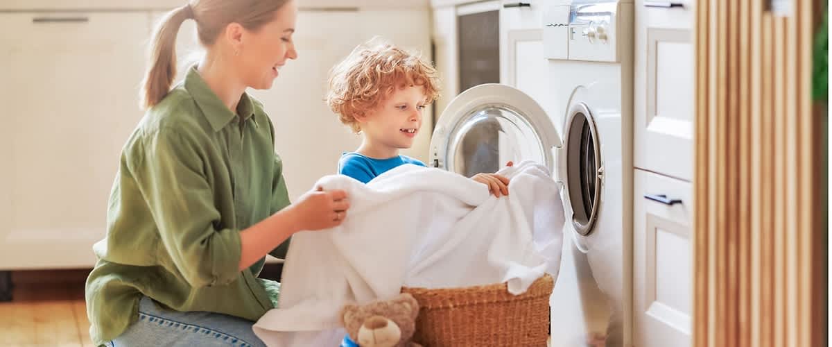 Zwei Personen, eine ältere sitzend im Schneidersitz und eine jüngere auf den Knien, legen zusammen weiße Wäsche aus einem Weidenkorb in die Frontlader-Waschmaschine der Küche; im Vordergrund liegt ein brauner Teddybär auf einem gestreiften Teppich, im Hintergrund Küchenschränke und Waschmaschinentür sichtbar