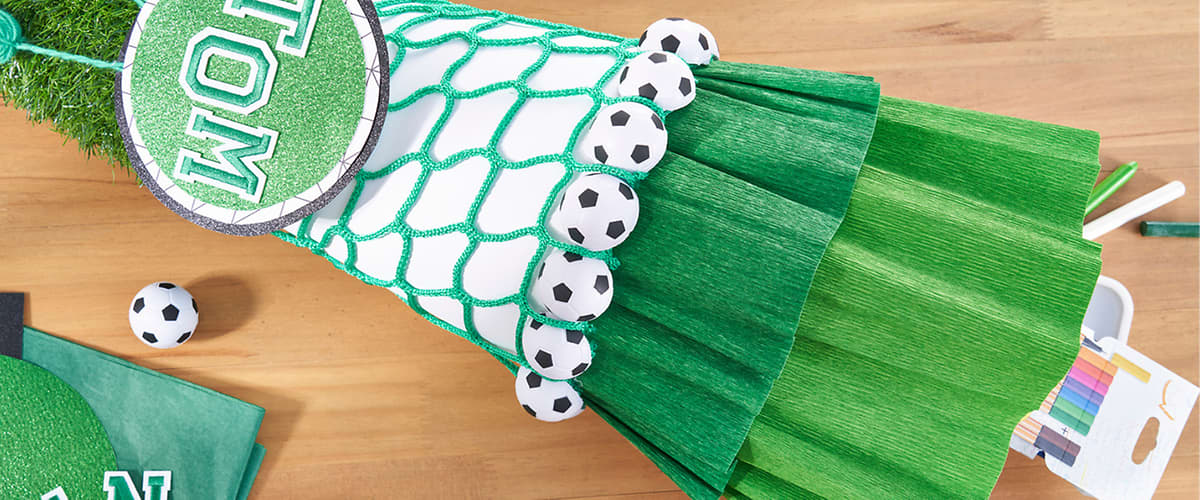 Mehrere grüne Krepppapierbahnen liegen gefaltet auf einem hellen Holztisch, oben links liegen fünf kleine Papiersoccerbälle als Deko, rechts neben dem Krepppapier befinden sich ein offenes Farbkasten-Set mit sichtbaren Buntstiften und ein paar grüne Papierstücke und Schaschlikspieße für Bastelarbeiten