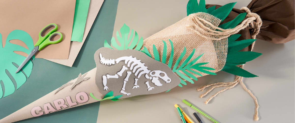 Eine Schultüte aus hellbeigem Papier mit grünen Papierblättern und Juteabschluss, seitlich beklebt mit einem grauen Dinosaurier-Skelett aus Papier und dem Namen „CARLO“ in rosa Buchstaben, umgeben von Bastelmaterialien wie Schere, Papierstreifen und Stiften