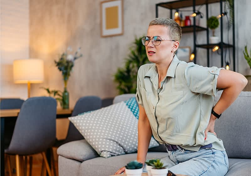Eine Person mittleren Alters sitzt seitlich auf einem grauen Sofa, stützt sich mit einer Hand auf die Sitzfläche und hält die andere Hand an den unteren Rücken, als hätte sie Rückenschmerzen, trägt kurzärmliges helles Hemd und Jeans und im Hintergrund ein Wohnraum mit Kissen, Stehlampe und Regal mit Dekorationen