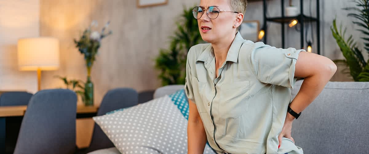 Eine Person mittleren Alters sitzt seitlich auf einem grauen Sofa, stützt sich mit einer Hand auf die Sitzfläche und hält die andere Hand an den unteren Rücken, als hätte sie Rückenschmerzen, trägt kurzärmliges helles Hemd und Jeans und im Hintergrund ein Wohnraum mit Kissen, Stehlampe und Regal mit Dekorationen