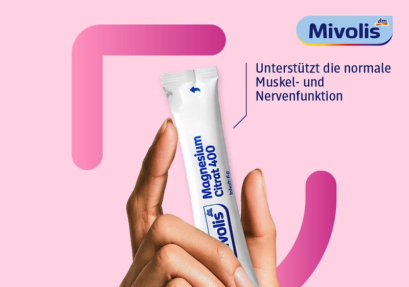 Hand hält ein weißes Einmal-Sachet mit blauem Aufdruck „Mivolis Magnesium Citrat 400“, rechts oben Firmenlogo „Mivolis“ und Text „Unterstützt die normale Muskel- und Nervenfunktion“ vor rosa Hintergrund mit dunkelrosa geometrischen Formen