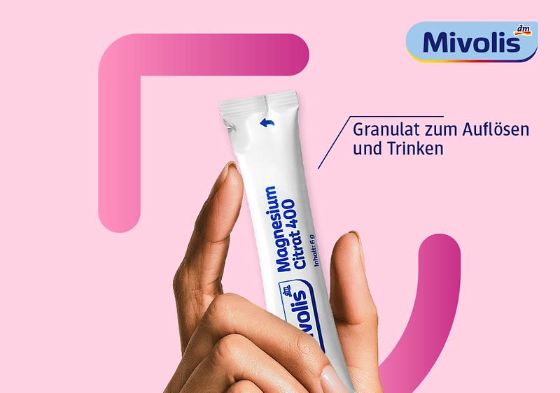 Hand hält ein weißes Einmal-Packstäbchen mit blauer Aufschrift „Mivolis Magnesium Citrat 400“ und blauem Markenlogo, daneben Text „Granulat zum Auflösen und Trinken“ und oben rechts das Mivolis-Logo vor rosafarbenem Hintergrund ohne Punkt am Satzende