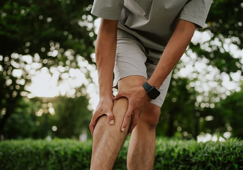 Ein Mann in Shorts hält sich mit beiden Händen die schmerzende rechte Schienbeinseite knapp unterhalb des Knies, im Hintergrund Bäume und Grünfläche