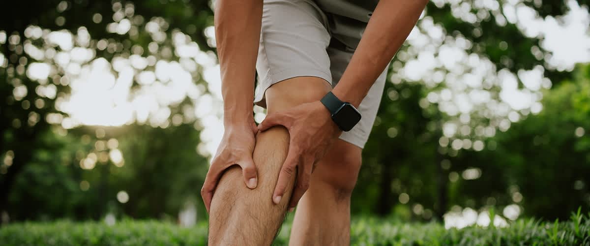 Ein Mann in Shorts hält sich mit beiden Händen die schmerzende rechte Schienbeinseite knapp unterhalb des Knies, im Hintergrund Bäume und Grünfläche