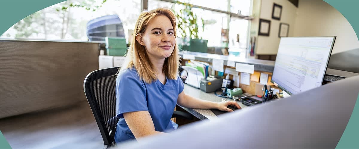 Eine am Empfang sitzende Mitarbeiterin in blauem Berufshemd arbeitet an einem Schreibtisch mit Computerbildschirm, Telefon und Büroorganisation, in hellem Empfangsbereich mit Fensterfront im Hintergrund