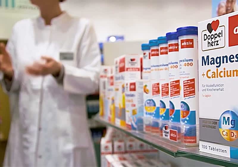 Regal mit Reihen von Nahrungsergänzungsmittelpackungen, im Vordergrund eine Dose „Doppelherz Magnesium + Calcium“ mit blauem und orangem Design und der Aufschrift „Magnesium + Calcium 100 Tabletten“, im Hintergrund eine leicht unscharf erkennbare Person in weißem Laborkittel mit Namensschild, die mit leicht erhobenen Händen steht