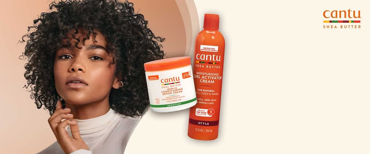 Frau mit krausem, schulterlangem Haar trägt ein weißes, langärmeliges Oberteil; vor ihr schwebend zwei orange-weiße Cantu-Produktverpackungen: links ein runder Tiegel mit der Aufschrift „Cantu Shea Butter Coconut Curling Cream“ und rechts eine hohe Dose mit der Aufschrift „Cantu Shea Butter Moisturising Curl Activation Cream“, rechts oben zusätzlich das Cantu-Logo mit dem Schriftzug „SHEA BUTTER“