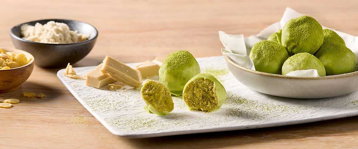 Drei grüne Matcha-Pralinen auf weißem Teller, eine halbiert und innen krümelig, daneben weiße Schokolade in Stücken und Matchapulver bestreut