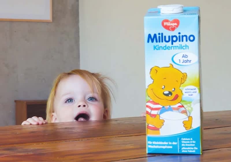 Kleiner Kindertisch: links hinter einer Holzplatte eine knieende Person, nur Hände am Tischrand sichtbar, rechts im Vordergrund eine blaue Milupino-Kindermilch-Packung mit gelbem Teddybär, Aufschrift „Milupino Kindermilch Ab 1 Jahr“ und Herstellerlogo Philippa, Packung steht auf dem Tisch